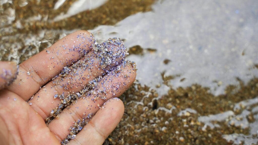 Microplastics clinging to fingers against a backdrop of the ocean, underscoring the perils they pose to marine ecosystems and the environment